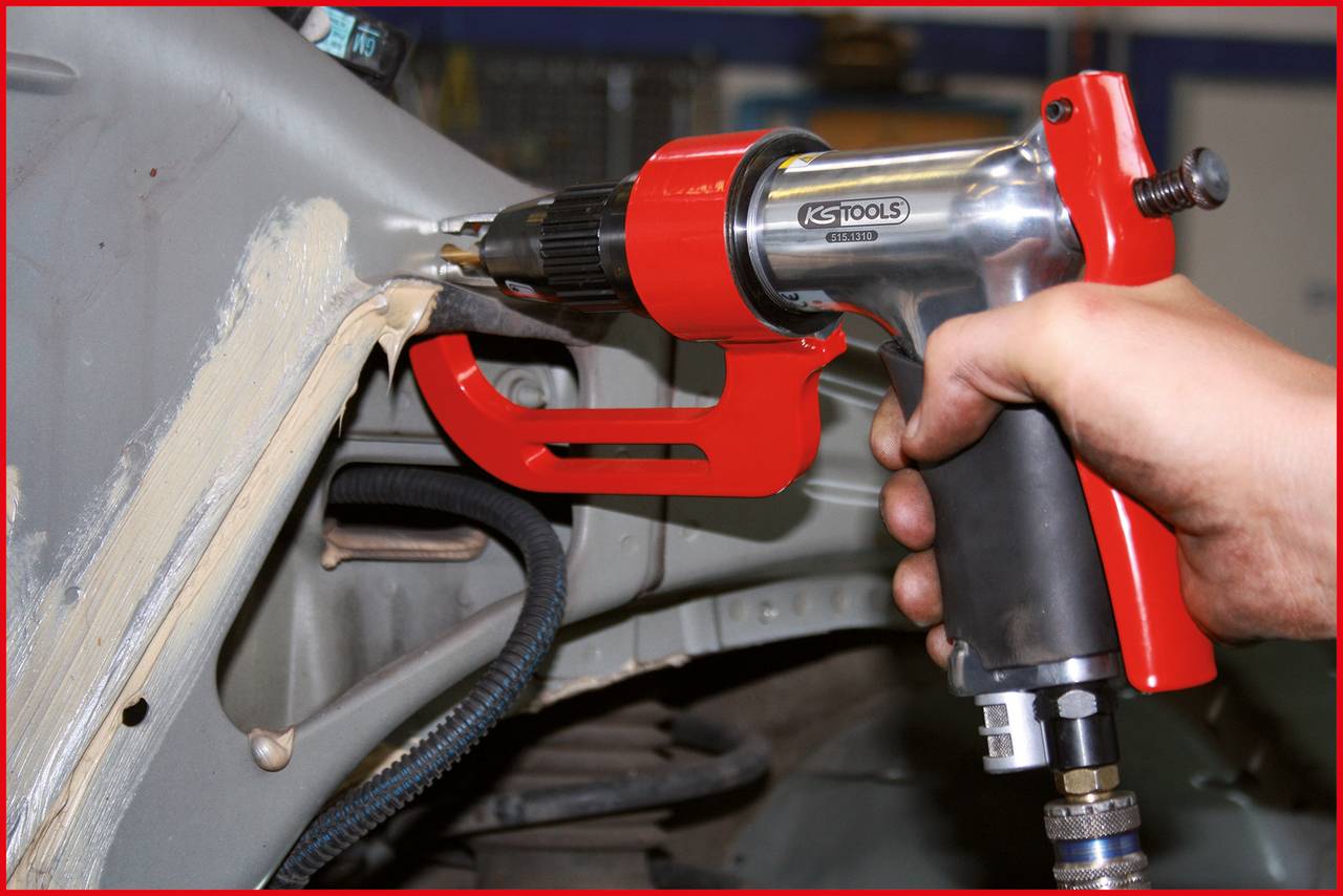 Eine Person benutzt einen Drucklufthammer, um Metall zu bearbeiten. Der Hammer hat einen roten Griff und wird an einem Auto verwendet.
