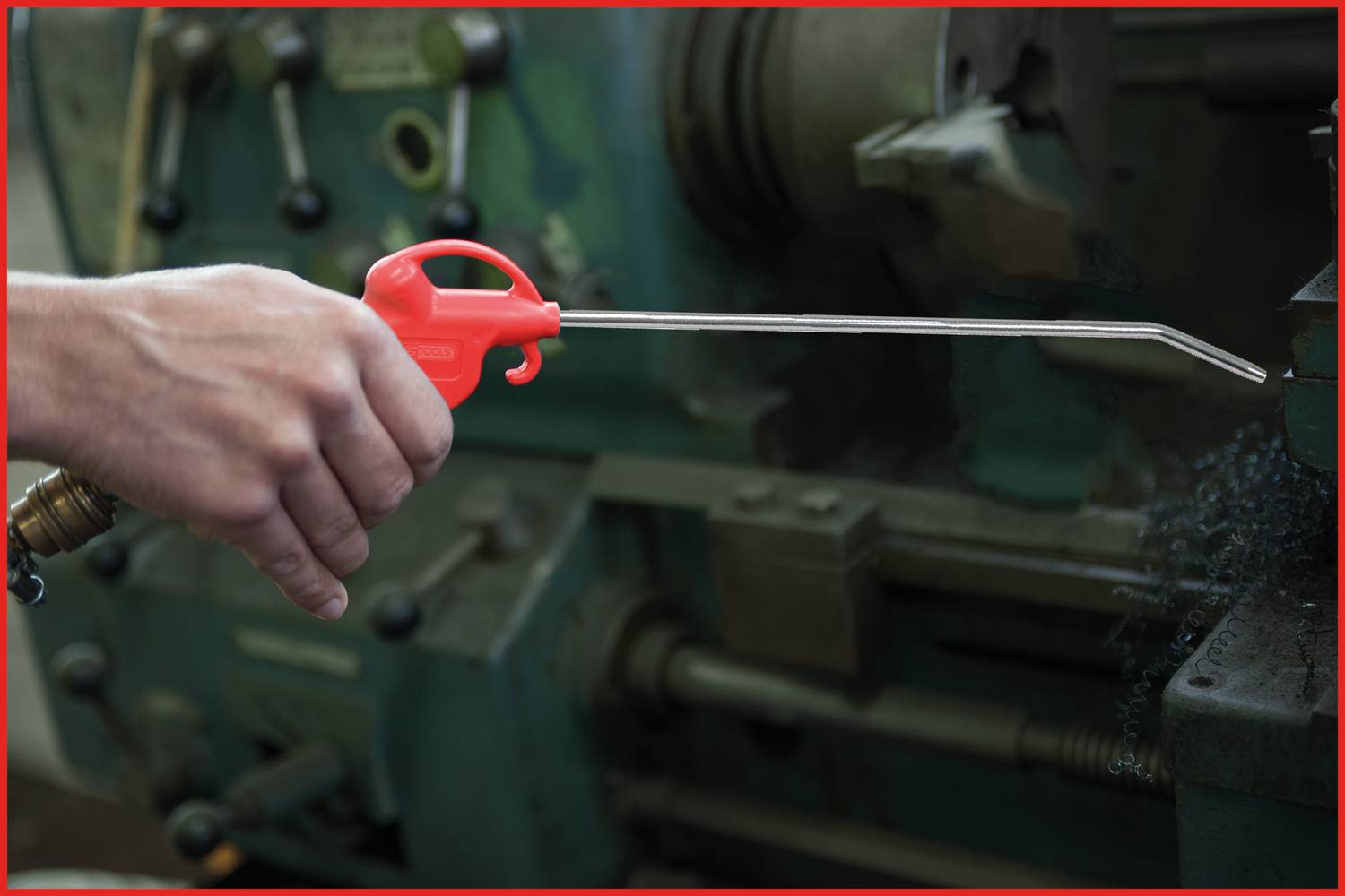 Eine Person hält ein Druckluftspray mit rotem Griff, um Metallspäne von einer Maschine zu entfernen. Die Maschine ist teilweise sichtbar.