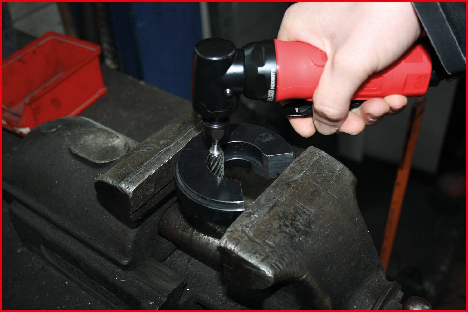 Eine Hand hält einen roten elektrischen Werkzeugschrauber, der auf einem Metallstück in einem Schraubstock arbeitet.