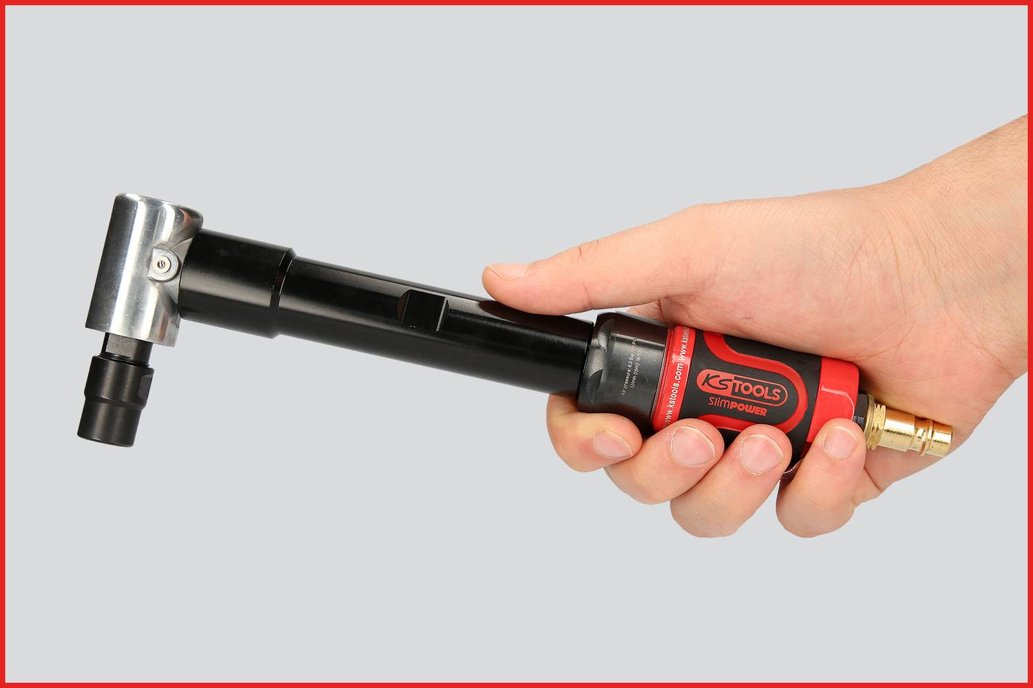 Eine Hand hält ein rot-schwarzes Werkzeug mit Metallkopf und Schlauchanschluss, möglicherweise ein Druckluftwerkzeug.