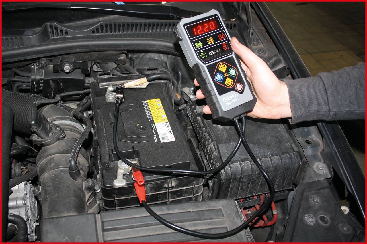 Ein Kfz-Techniker verwendet ein digitales Batterietestgerät zur Überprüfung der Autobatterie. Das Display zeigt '12,20'.