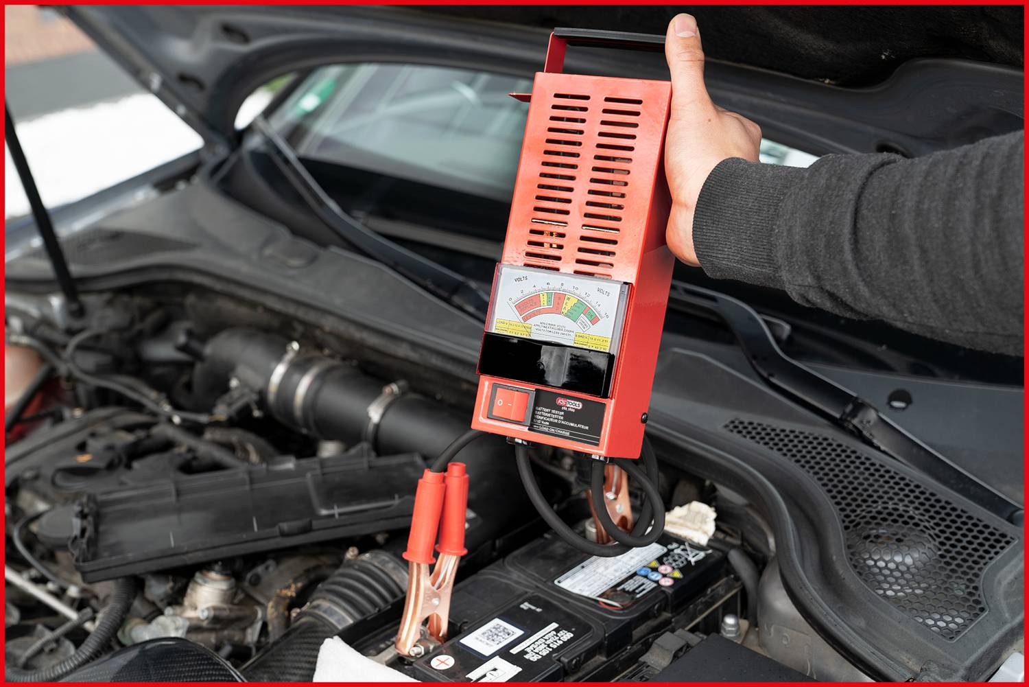 Eine Person testet die Autobatterie eines offenen Motorraums mit einem Batterietester. Das Gerät zeigt den Ladestand an.
