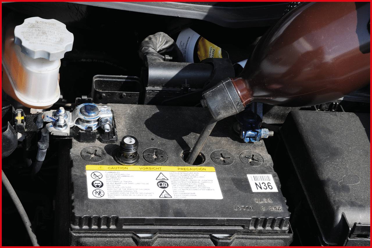 Batterie eines Fahrzeugs mit geöffnetem Deckel, daneben eine Plastikflasche, die Flüssigkeit nachfüllt. Im Hintergrund ein Behälter mit Flüssigkeit.