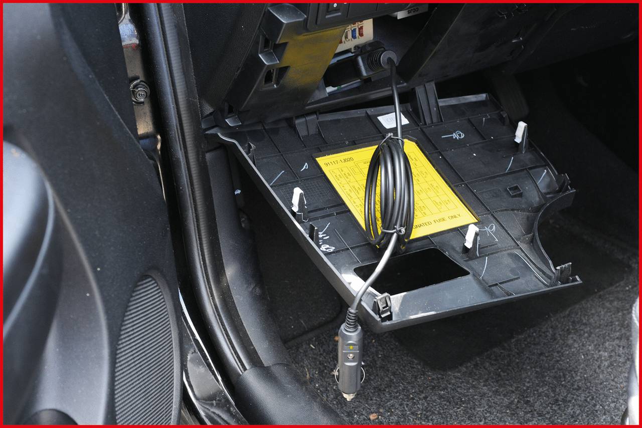 Geöffnetes Handschuhfach eines Autos mit herausgenommenem Boden, ein gelbes Warnschild ist sichtbar, Kabel hängt heraus.