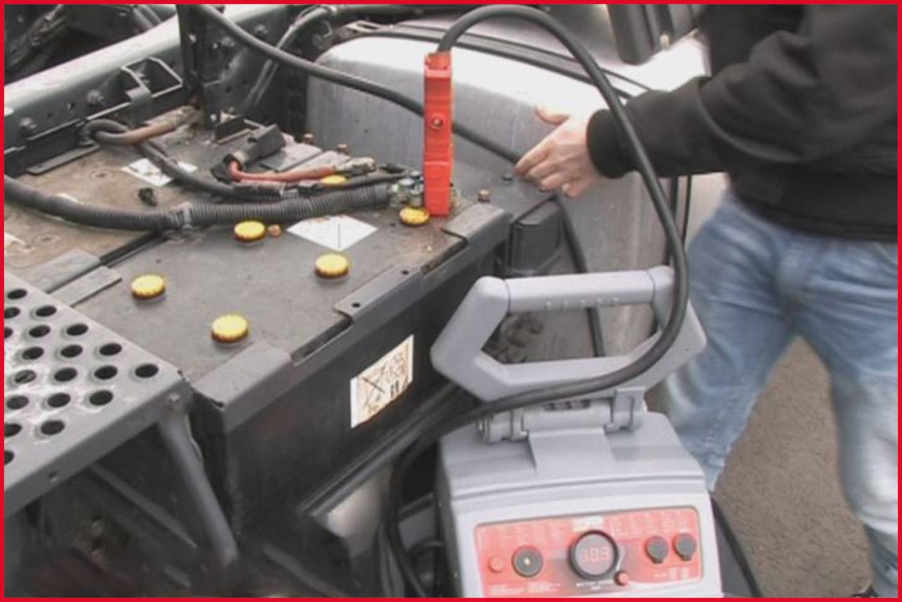 Person lädt Autobatterie mit einem Starthilfegerät auf. Rotes Kabel ist angeschlossen. Hände sind sichtbar, Fokus auf der Batterie.