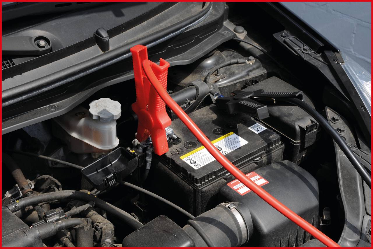 Eine Autobatterie wird mit Starterkabeln überbrückt, rote Klemme am Pluspol. Der Motorraum ist geöffnet und sichtbar.