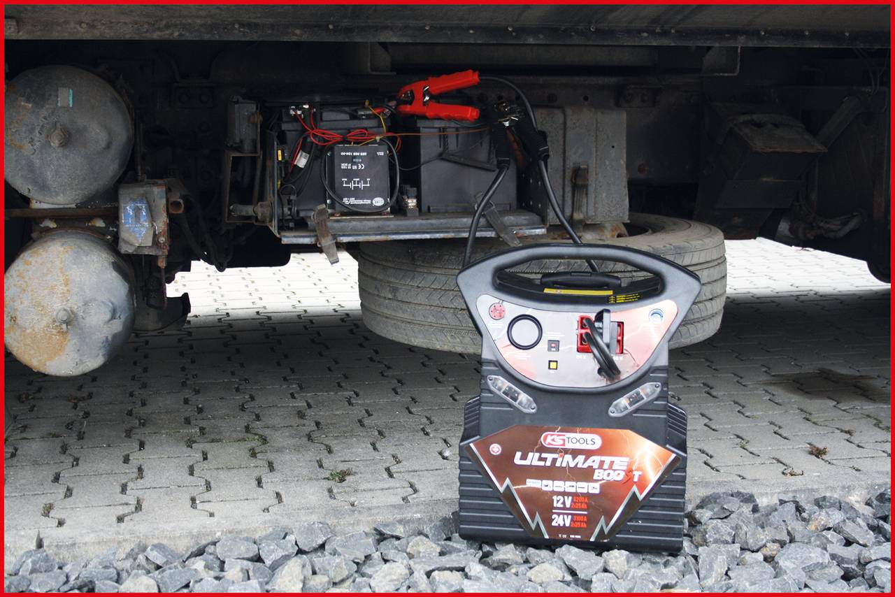 Ein Batterieladegerät ist an eine Autobatterie angeschlossen, um sie aufzuladen. Der Lkw steht auf einem gepflasterten Untergrund.