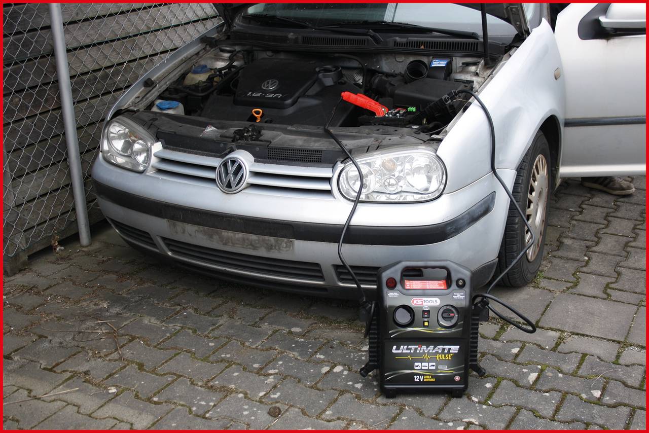 Ein Auto mit offener Motorhaube wird mit einem Starthilfegerät überbrückt. Das Gerät ist mit dem Motor verbunden und auf dem Boden platziert.