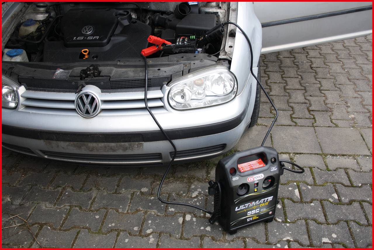 Auto wird mit einem tragbaren Batterie-Booster aufgeladen. Das Fahrzeug steht auf Kopfsteinpflaster, die Motorhaube ist geöffnet.