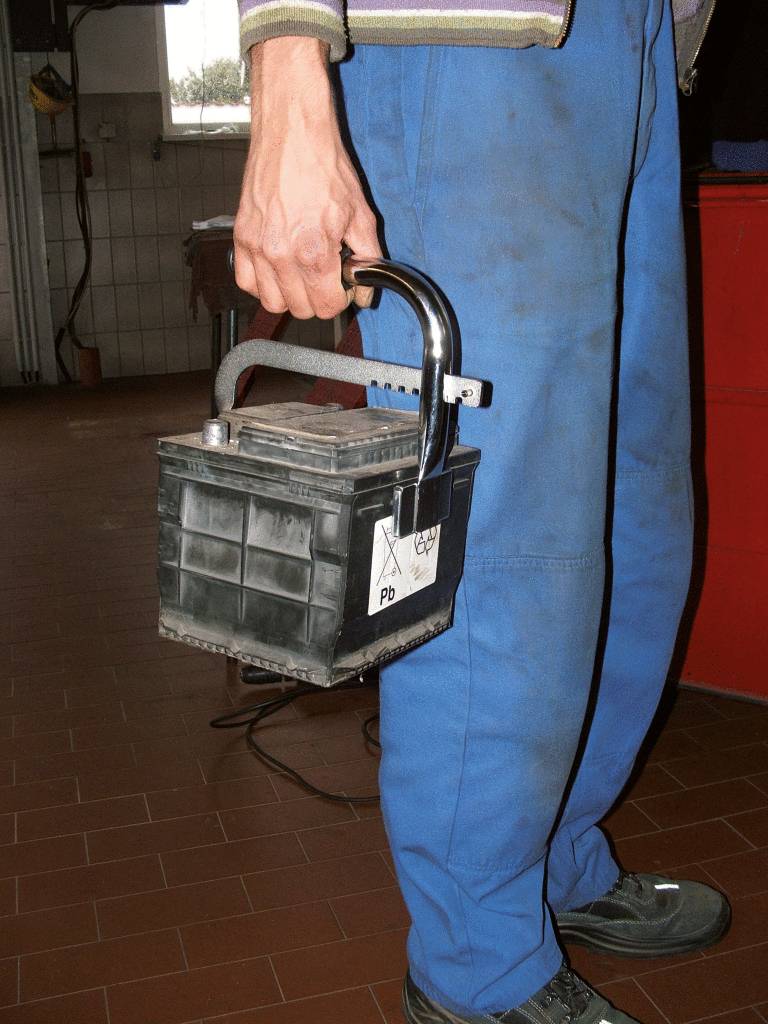 Eine Person in blauer Arbeitskleidung trägt eine Autobatterie mit einem Griff. Sie befindet sich in einer Werkstattumgebung.