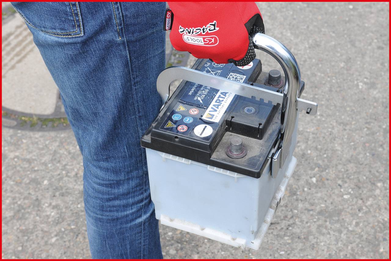 Eine Person in blauen Jeans hält eine Auto-Batterie mit der Hand. Die Person trägt rote Arbeitshandschuhe.