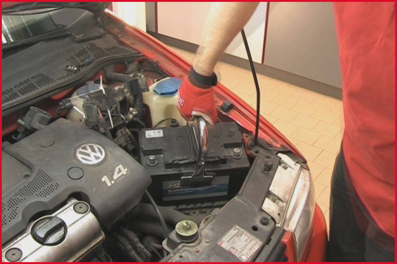 Ein Mechaniker überprüft die Autobatterie in einem roten Fahrzeug. Eine Hand hält ein Diagnosewerkzeug auf der Batterie.