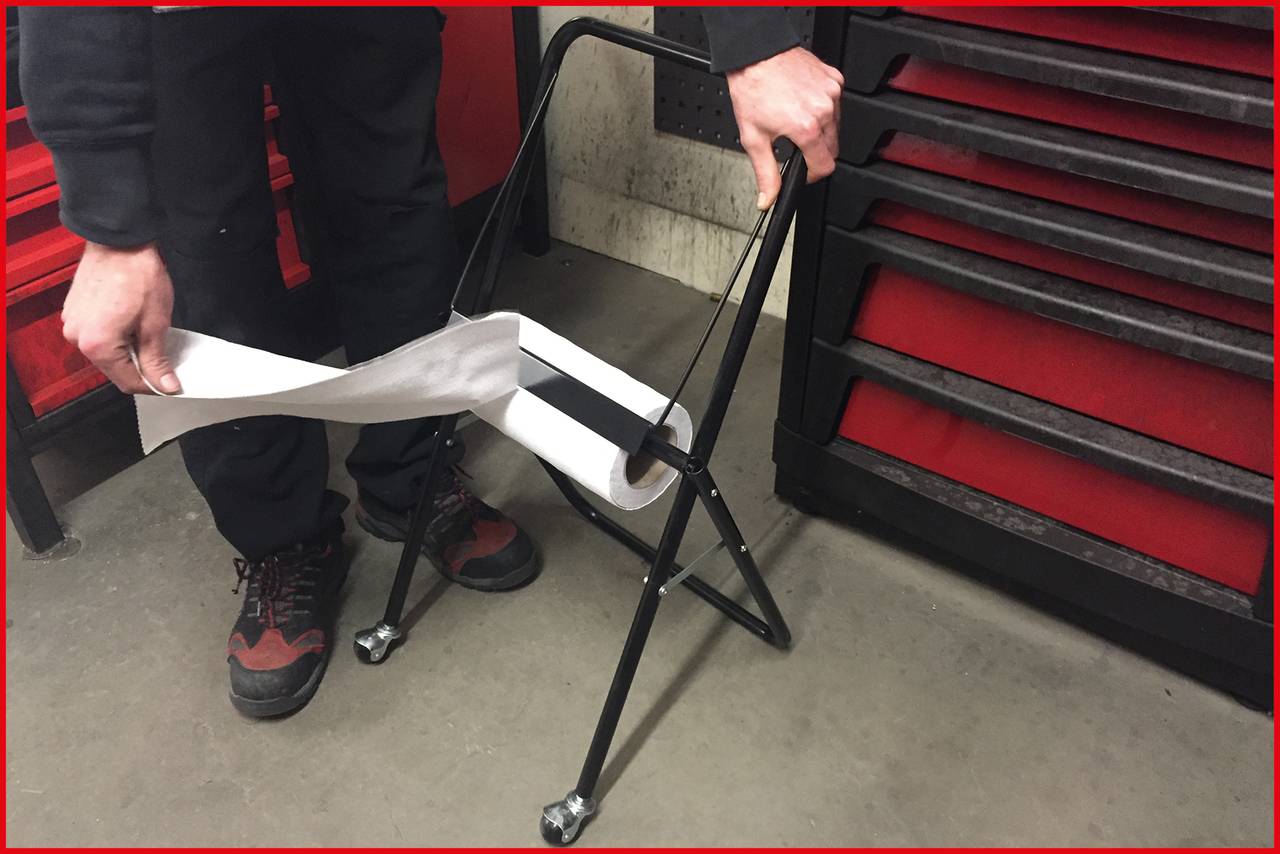 Eine Person zieht Papier von einem schwarzen Metallständer mit Rädern ab. Links steht eine rote Werkzeugkiste.