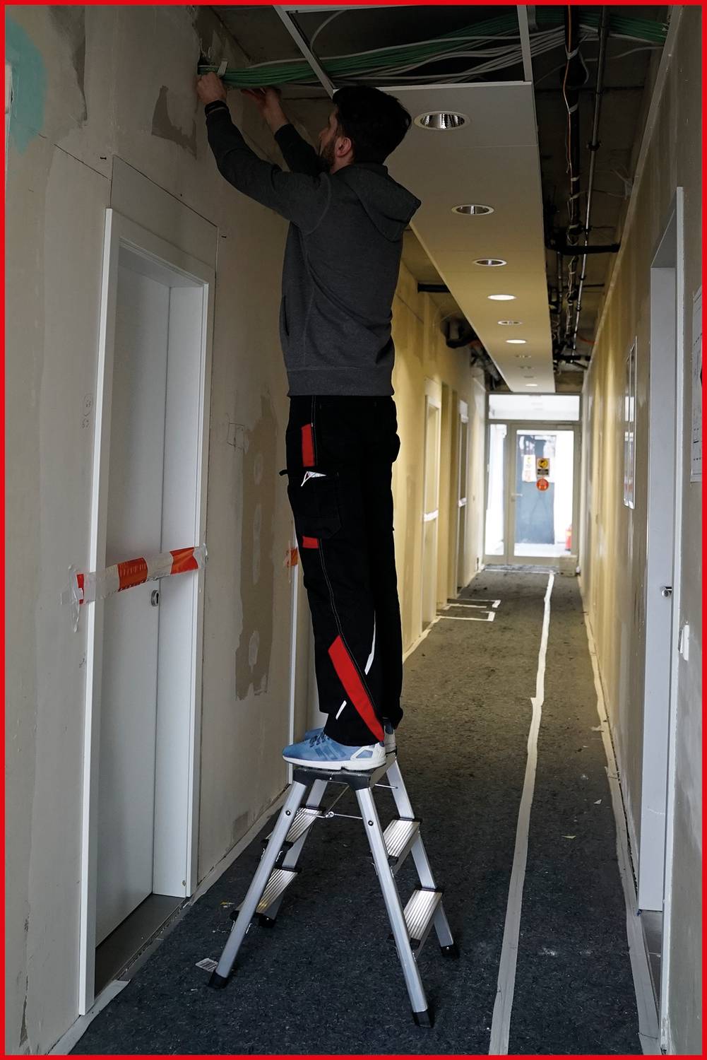 Ein Mann steht auf einer Leiter und arbeitet an der Decke in einem langen Flur. Teile der Wand sind unvollständig verputzt.