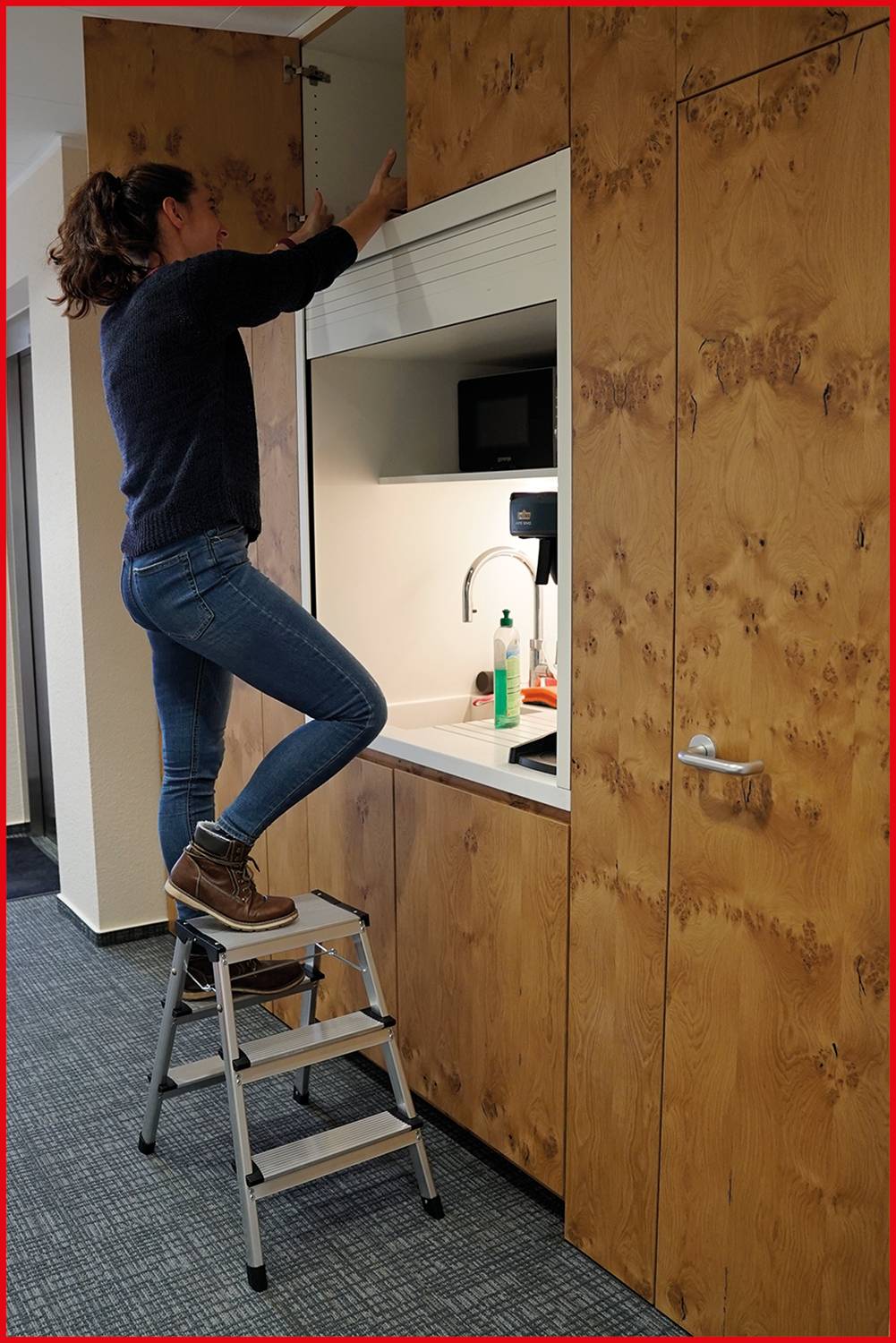 Eine Person steht auf einer kleinen Leiter und öffnet einen Schrank über einer Küchenzeile.