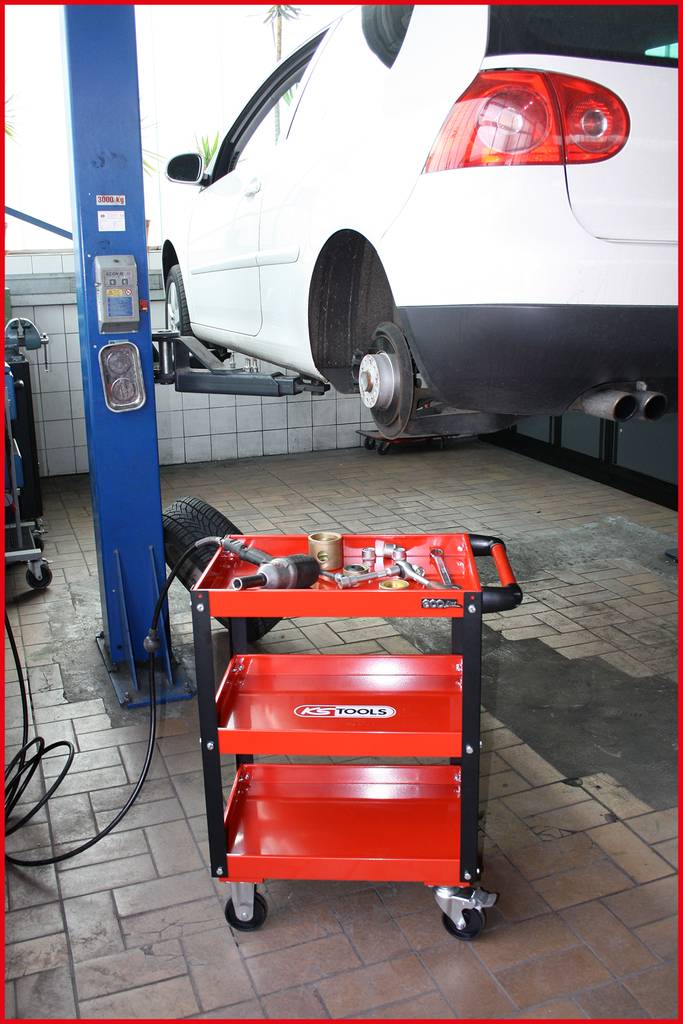 Ein weißes Auto wird in einer Werkstatt auf einer Hebebühne angehoben. Ein roter Werkzeugwagen steht daneben mit verschiedenen Werkzeugen.