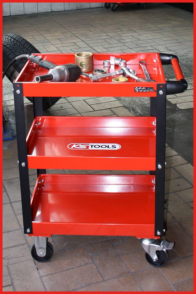 Roter Werkstattwagen mit drei Ablageflächen, darauf verschiedene Werkzeuge wie eine Bohrmaschine, in einer Autowerkstatt.