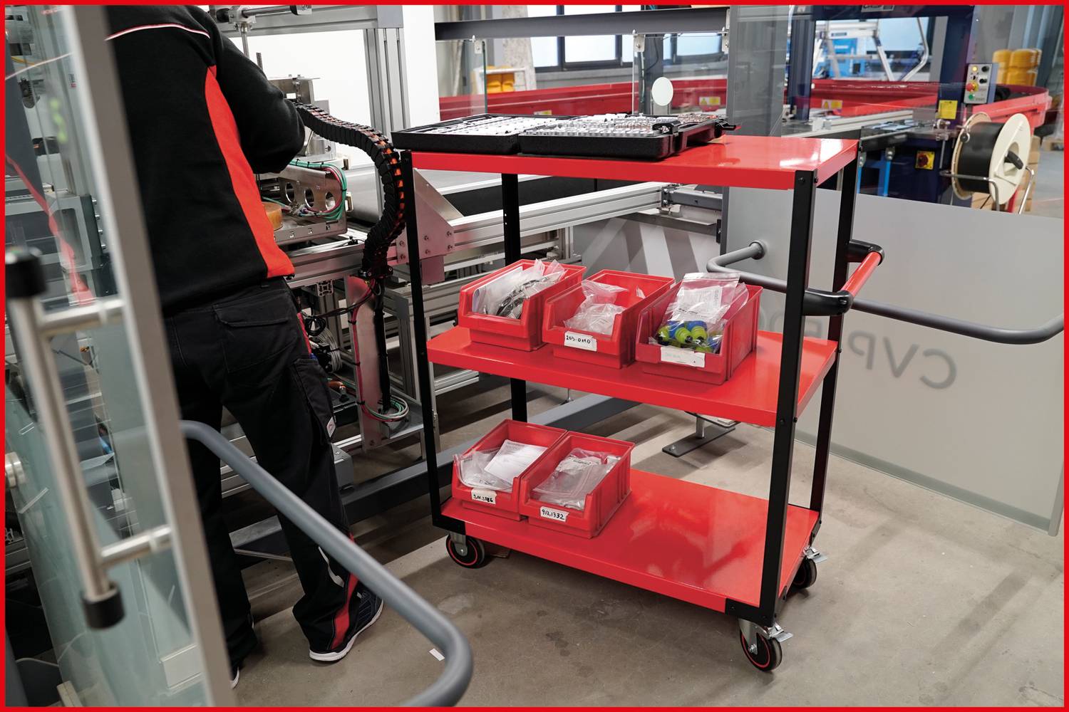 Eine Person in Arbeitskleidung steht an einer Maschine in einer Werkstatt. Ein roter Rollwagen mit verschiedenen Kleinteilen steht daneben.