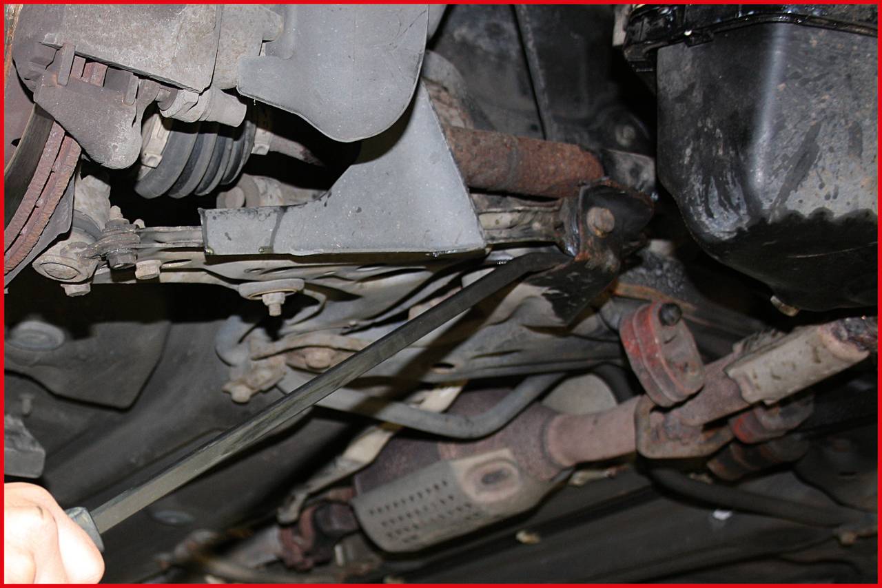 Ein Mechaniker zieht an einem Hebel unter einem Auto, um die Mechanik zu justieren. Sichtbar sind Fahrwerksteile und Auspuffsystem.