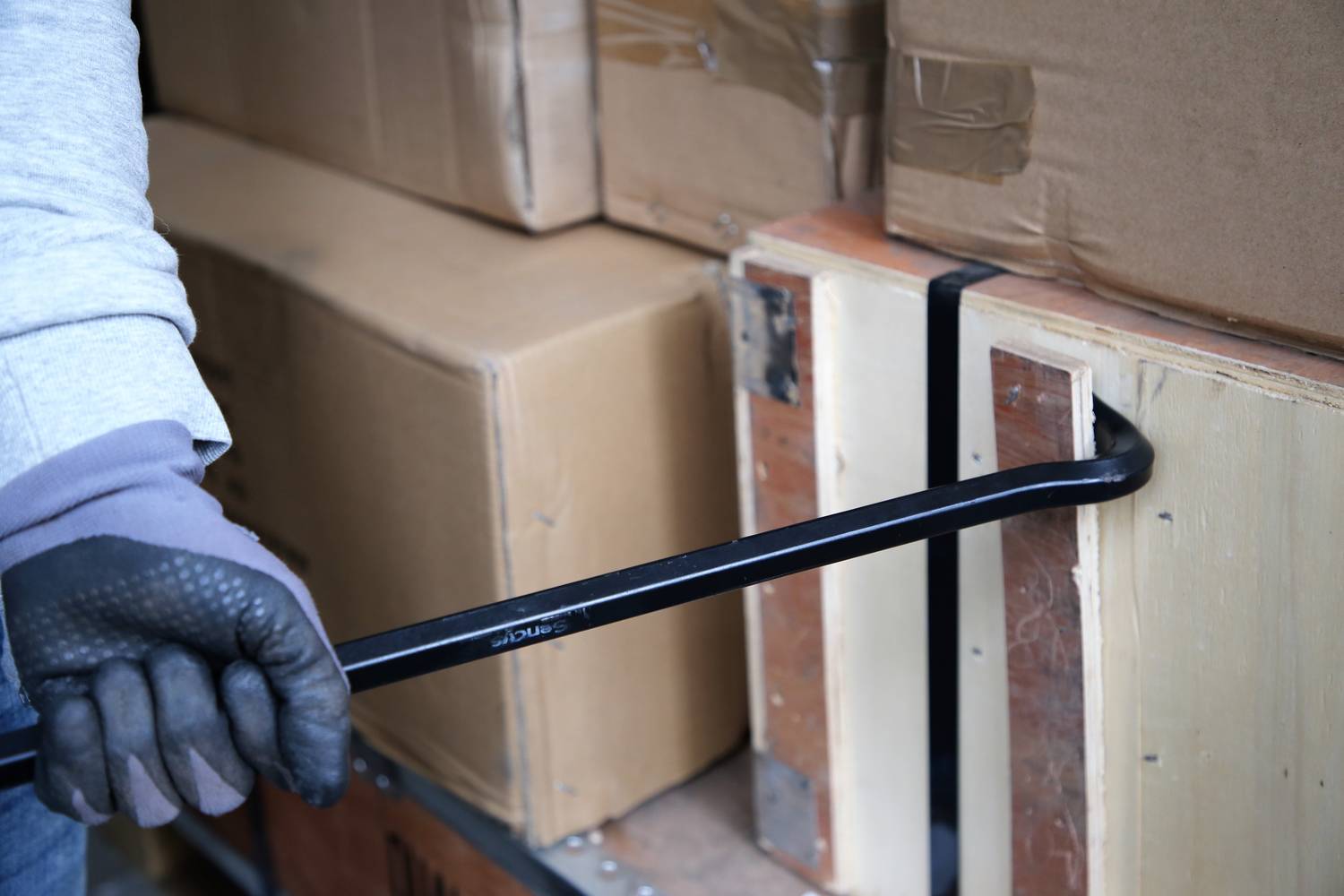 Eine Person hebelt mit einer Brechstange eine Kiste auf. Die Kiste ist aus Holz, und um sie herum sind andere Kartons gestapelt.