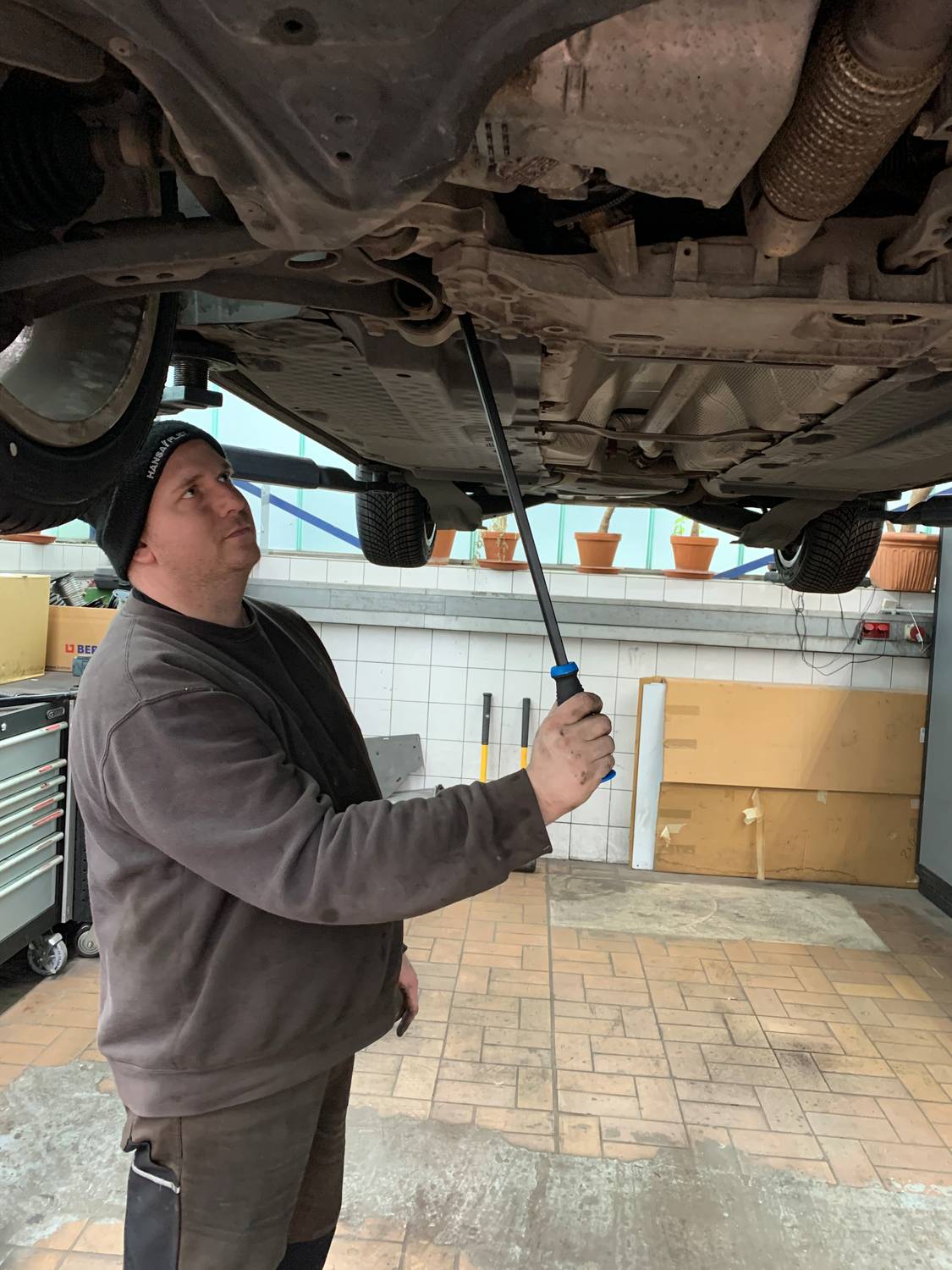 Eine Person inspiziert ein Fahrzeug, das aufgebockt ist. Sie nutzt eine Taschenlampe, um den Unterboden im Autowerkstattbereich zu überprüfen.