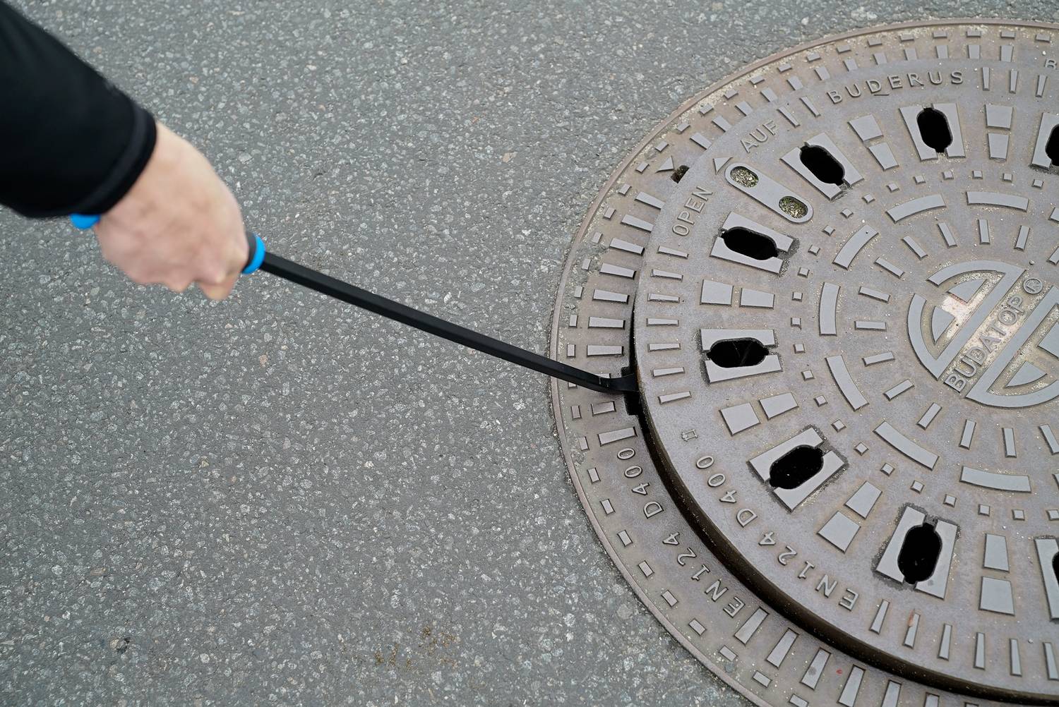 'Hand hebt einen Kanaldeckel mit Werkzeug' zeigt nahes Bild einer Straße.