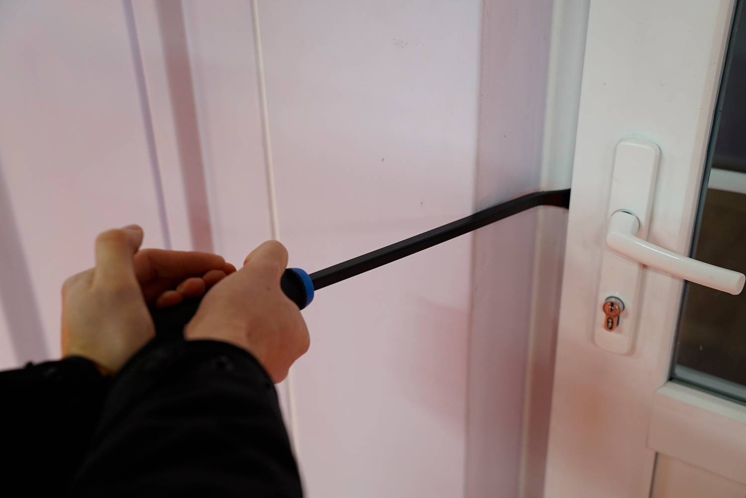Eine Person versucht, mit einem Brecheisen die Tür eines Hauses aufzubrechen. Die Tür ist weiß und der Hintergrund ist unscharf.