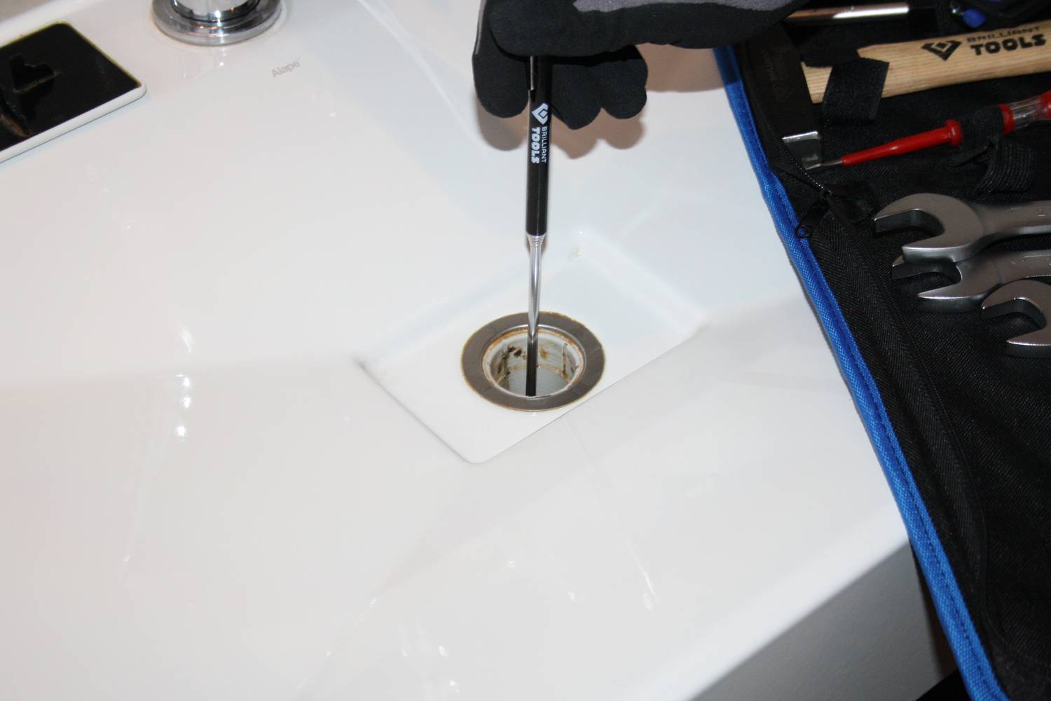Eine Person arbeitet mit Werkzeug an einem Waschbeckenabfluss. Mehrere Werkzeuge liegen auf einer Stofftasche neben dem Waschbecken.