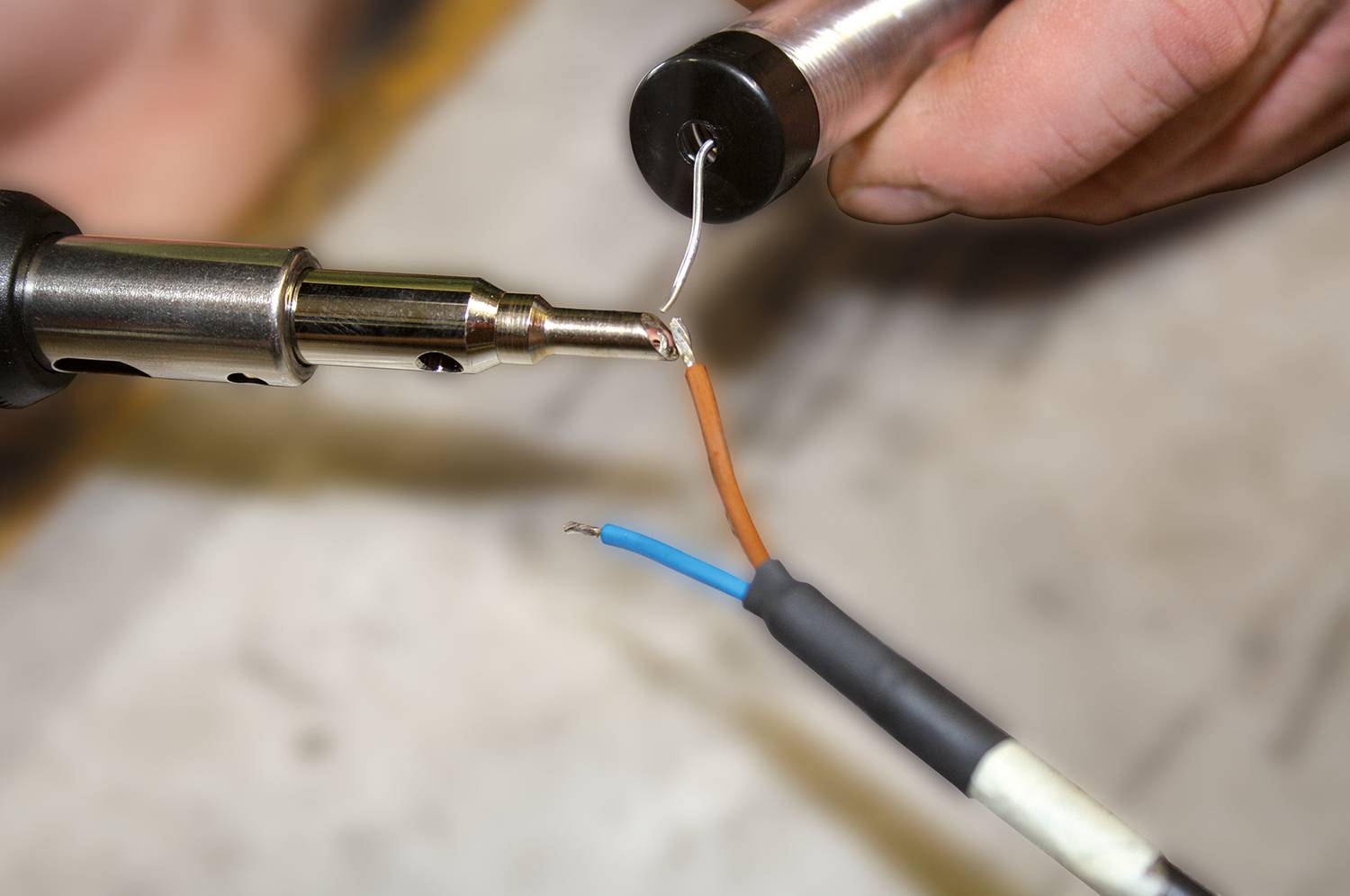 Eine Nahaufnahme von zwei Händen, die mit einem Lötkolben einen schwarzen Stecker an Drahtkabel mit brauner und blauer Isolierung löten.