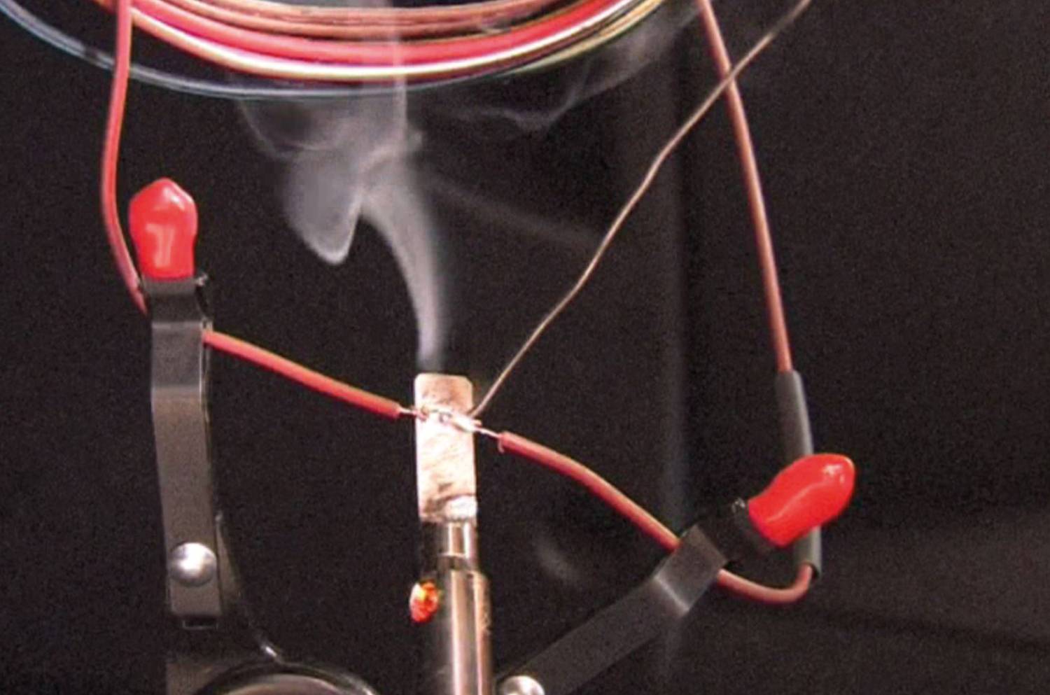 Lötkolben erhitzt ein Metallstück, das zwischen zwei roten Klemmen gehalten wird. Rauch steigt auf, während die Drähte verbunden werden.