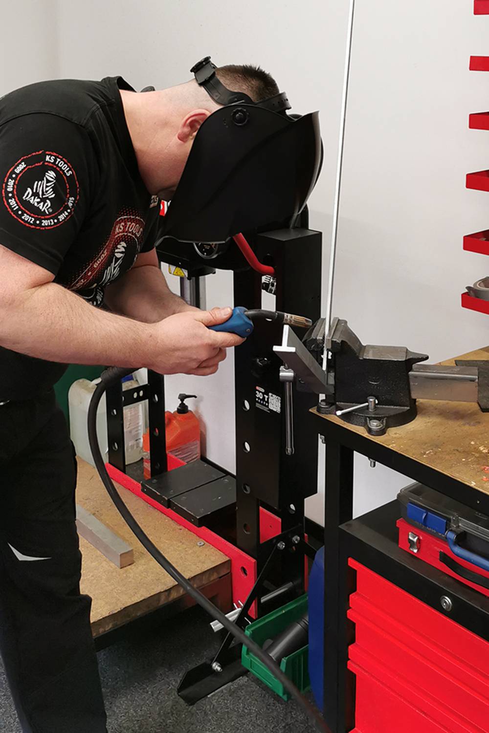 Ein Mann mit Schweißhelm arbeitet an einem Metallstück in einer Werkstatt. Er verwendet ein Schweißgerät neben einem Schraubstock.