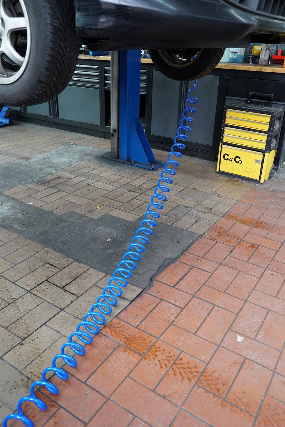 Auto in Werkstatt auf einer Hebebühne mit einem blauen, spiralförmigen Druckluftschlauch am Boden. Reifen teilweise sichtbar.