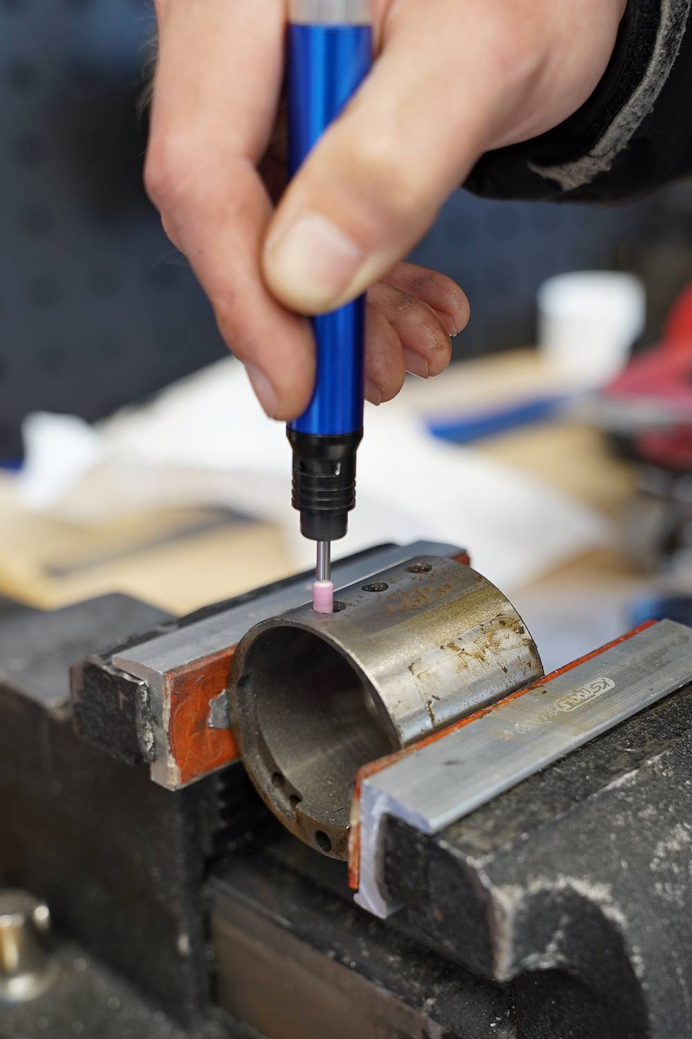 Eine Hand bearbeitet mit einem blauen Rotationswerkzeug ein Metallrohr, das in einem Schraubstock befestigt ist.