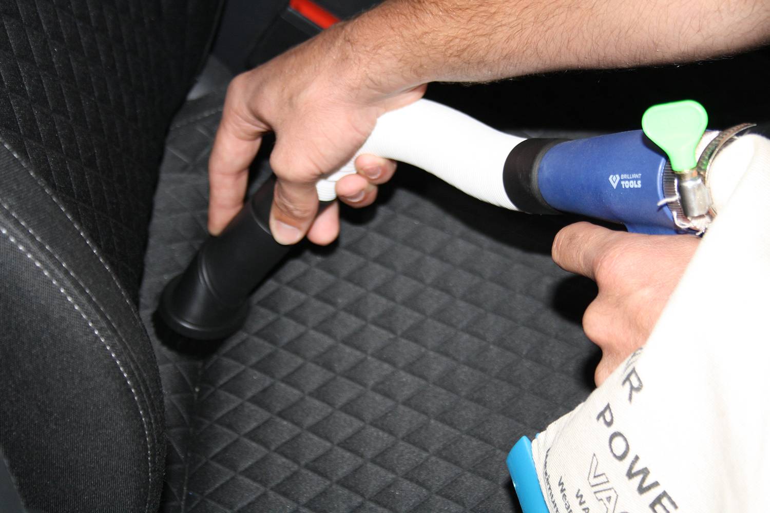 Eine Person reinigt einen Autositz mit einem Staubsauger, wobei der Fokus auf der Polsterdüse liegt.
