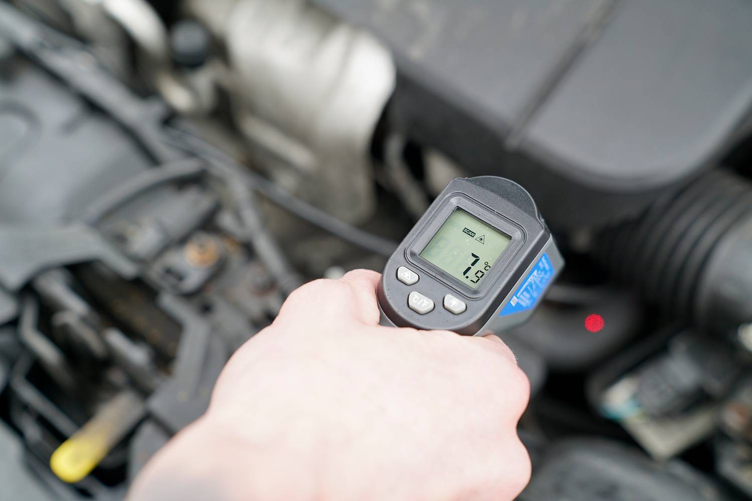 Eine Hand hält ein Infrarot-Thermometer über einem geöffneten Automotor und zeigt eine Temperatur von 51 Grad Celsius an.