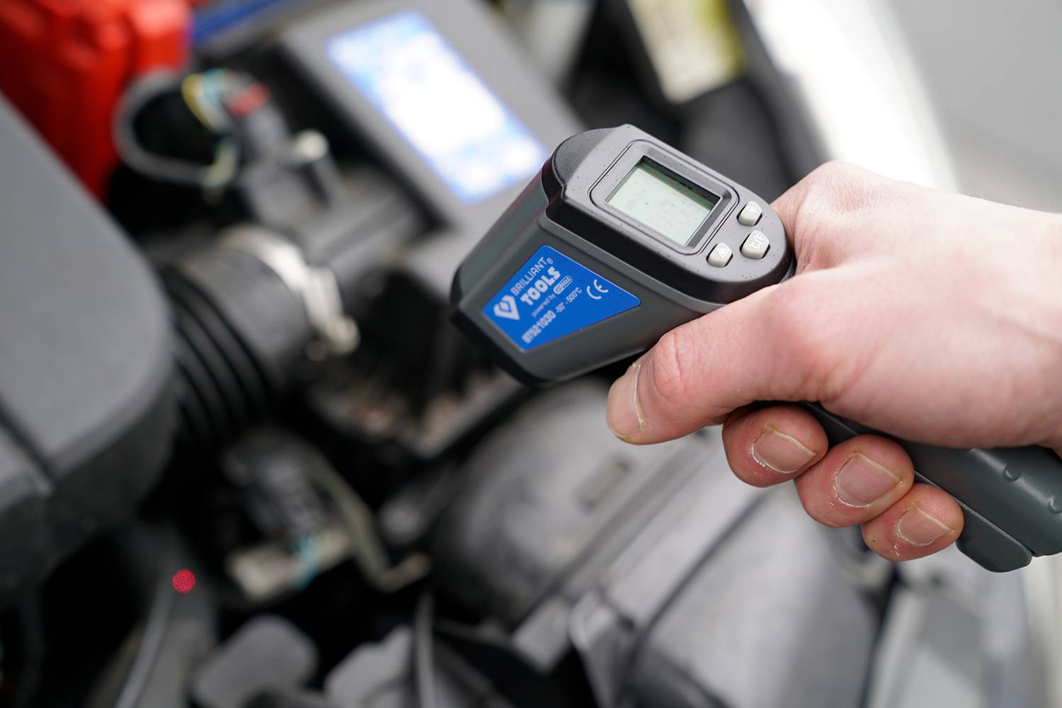 Hand hält ein Infrarot-Thermometer über einem Autoteil, misst die Temperatur des Motors. Anzeige zeigt Digitalwert.