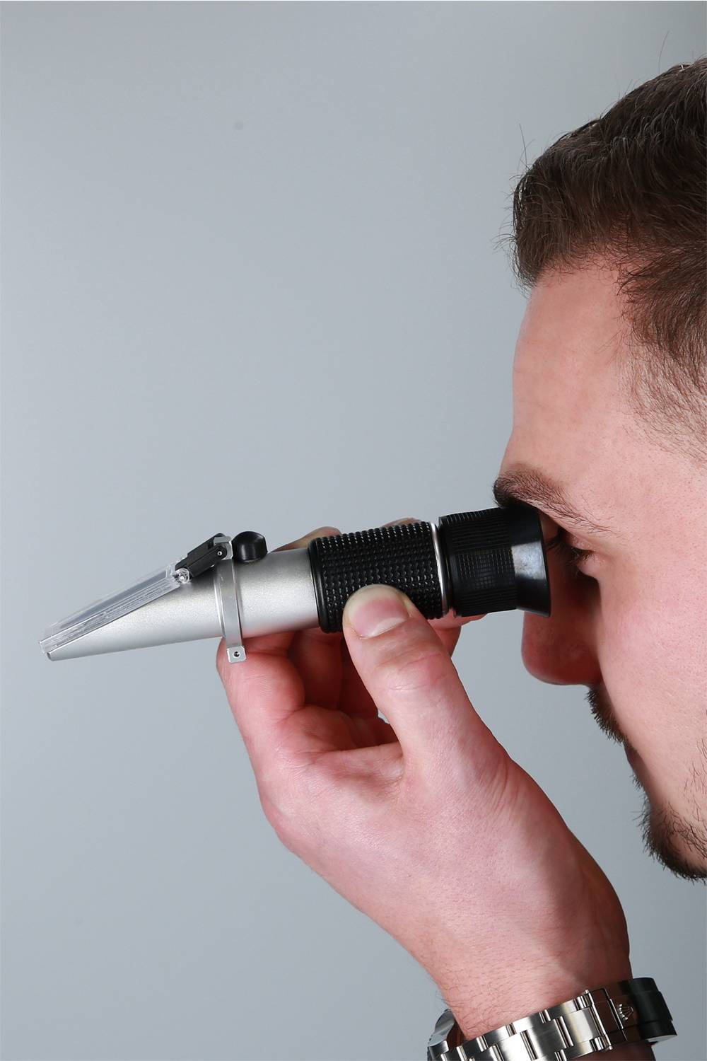 Ein Mann blickt durch ein silbernes Refraktometer, das er mit der rechten Hand hält.