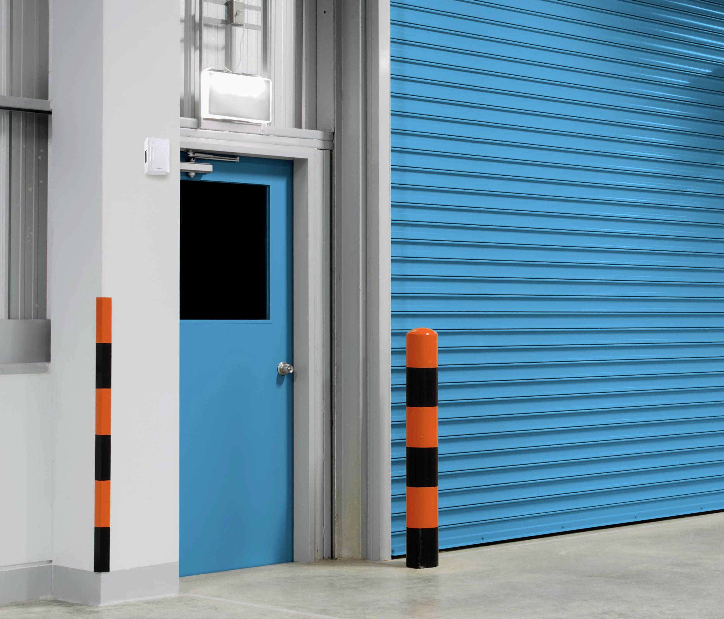 Eine blaue Tür neben einem großen, geschlossenen, blauen Rolltor in einem industriellen Gebäude. Im Vordergrund stehen orange-schwarze Pfosten.