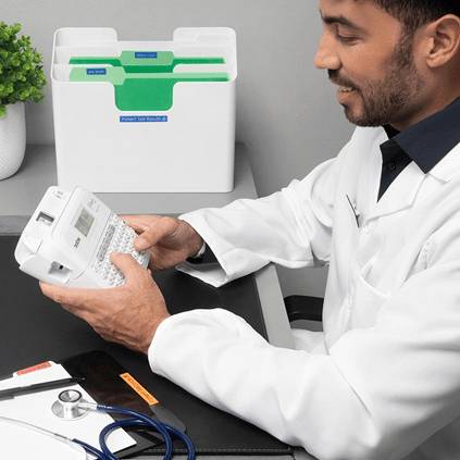 Ein Mann in einem weißen Kittel sitzt an einem Tisch und betrachtet ein Gerät. Ein grüner Ablagekorb steht im Hintergrund.