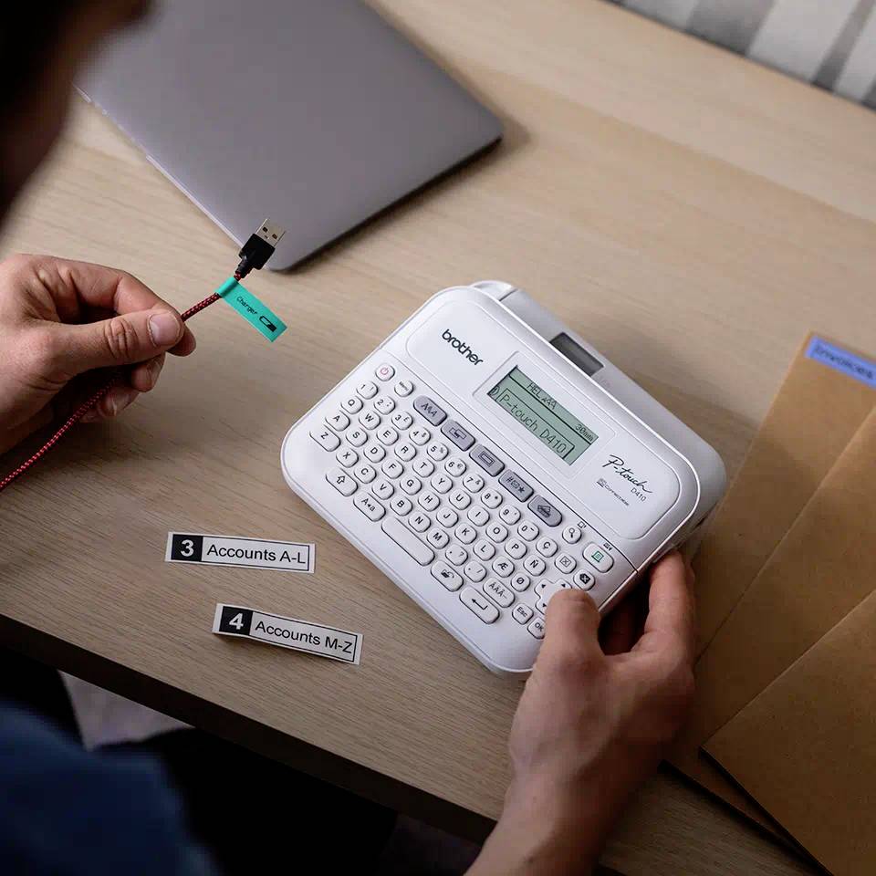 Eine Person verwendet ein Beschriftungsgerät, um Etiketten zu drucken. Ein Laptop und Umschläge liegen auf dem Tisch.