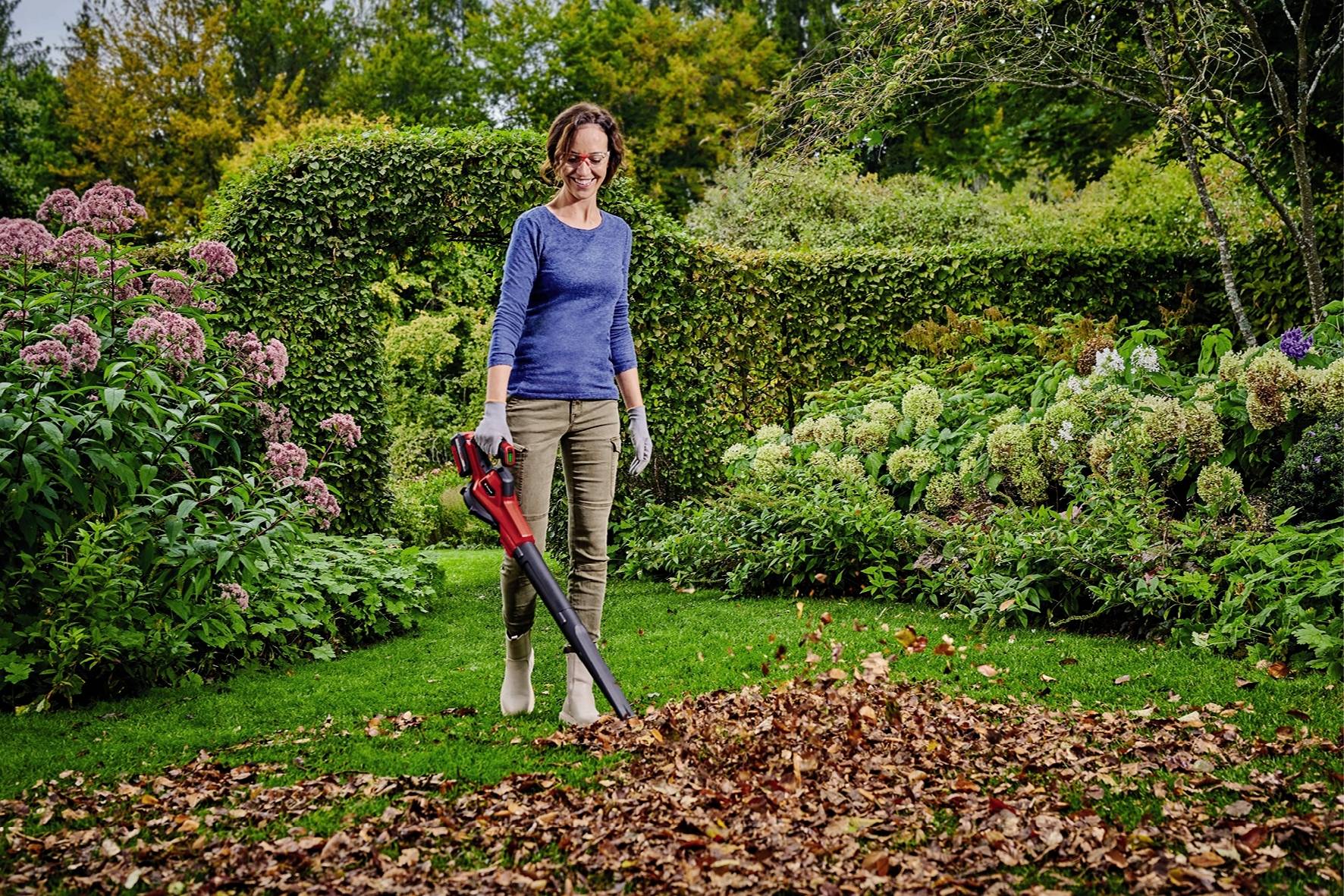 Eine Frau steht im Garten und bläst mit einem Laubbläser Laub zusammen. Sie trägt Gartenhandschuhe. Im Hintergrund sind Büsche und Bäume.