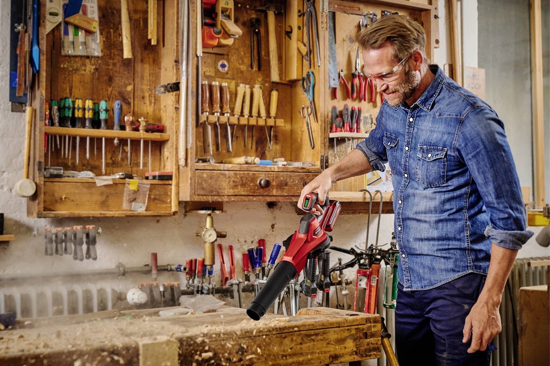 Mann in Werkstatt benutzt einen Handstaubsauger, um Holzspäne von einer Werkbank zu entfernen. Im Hintergrund sind Werkzeuge an der Wand.