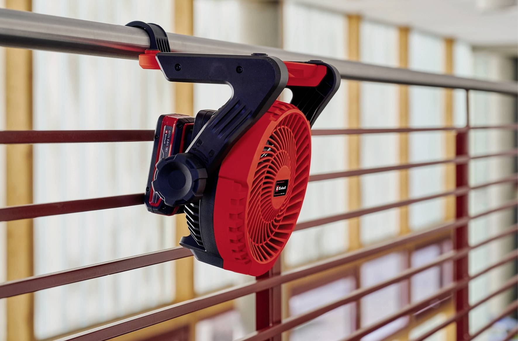 Ein roter, tragbarer Ventilator, der an einem Geländer angebracht ist. Im Hintergrund sind Fenster mit hellen Gardinen zu sehen.