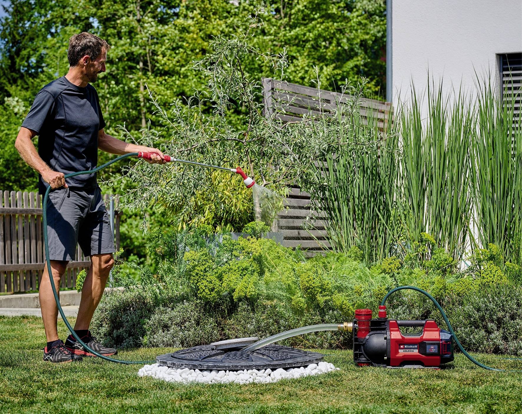 Ein Mann bewässert mit einem Gartenschlauch Pflanzen in einem Garten. Neben ihm steht eine rote Wasserpumpe. Im Hintergrund sind Bäume.