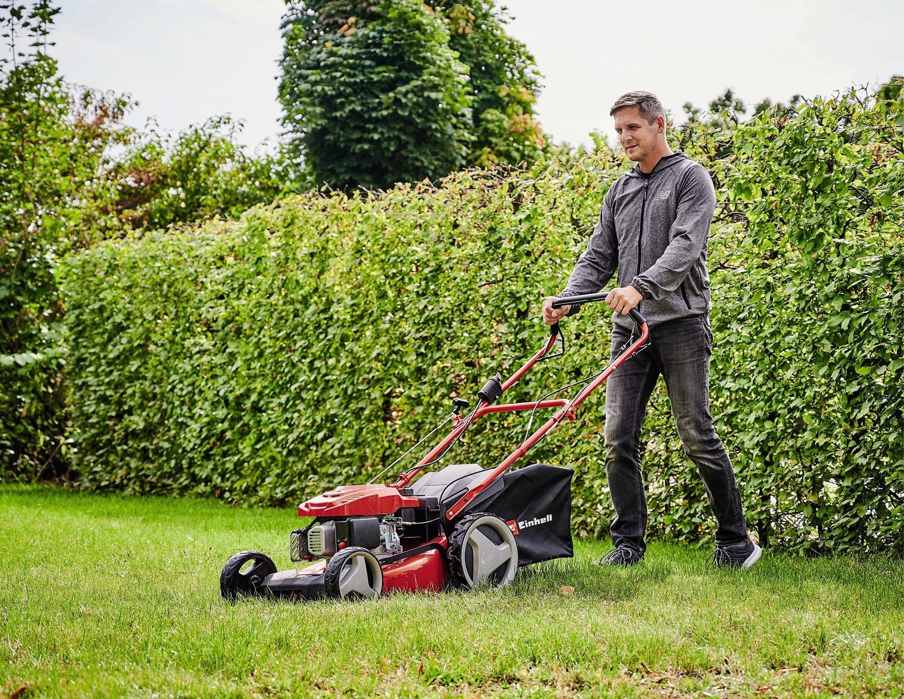 Eine Person mäht den Rasen im Garten mit einem roten Rasenmäher. Im Hintergrund ist eine hohe Hecke und einige Bäume zu sehen.