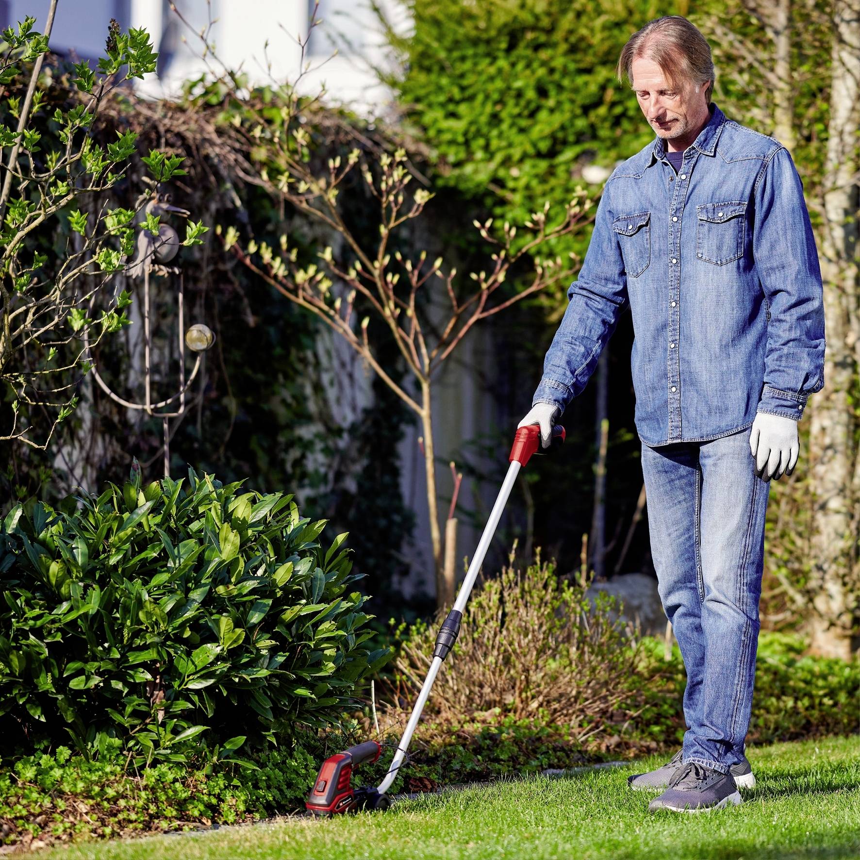 Eine Person in Jeanskleidung und Handschuhen trimmt den Rasen um einen Gartenbusch mit einem elektrischen Rasentrimmer.