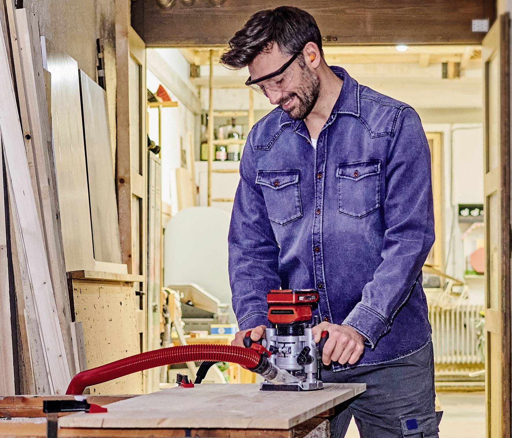Ein Mann in Arbeitskleidung benutzt eine Holzfräse in einer Werkstatt. Schutzbrille tragend, konzentriert er sich auf die Holzplatte.