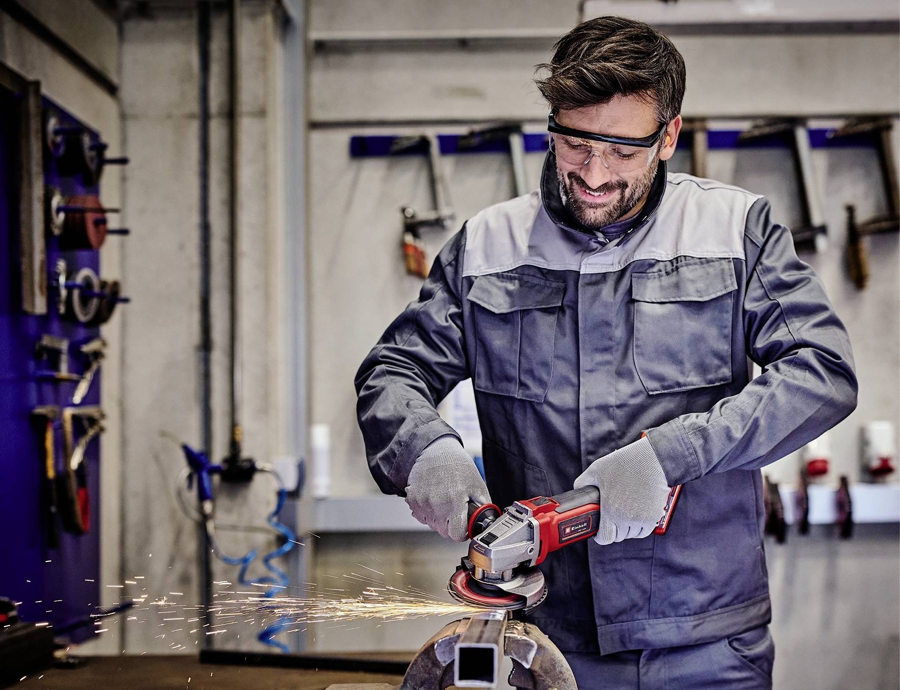 Ein Mann in Arbeitskleidung schleift Metall mit einem Winkelschleifer in einer Werkstatt. Funken fliegen. Schutzbrille und Handschuhe werden getragen.