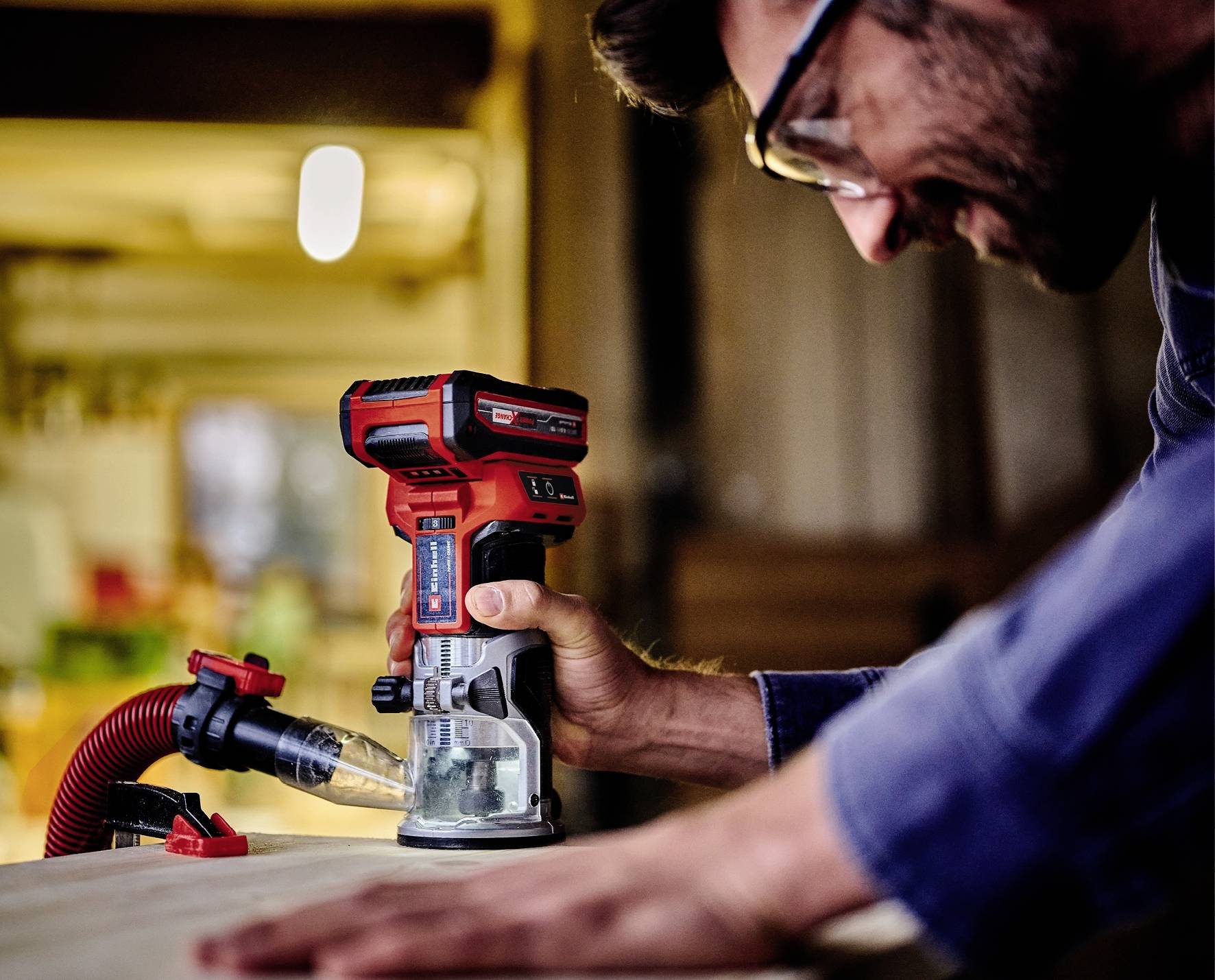 Ein Mann benutzt eine rote Handoberfräse auf einem Holzstück in einer Werkstatt. Er trägt eine Schutzbrille und konzentriert sich auf die Arbeit.
