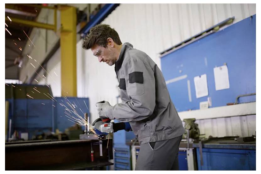 Eine Person in Schutzausrüstung verwendet einen Schleifer und erzeugt Funken in einer Industriewerkstatt mit blauen und weißen Wänden im Hintergrund.